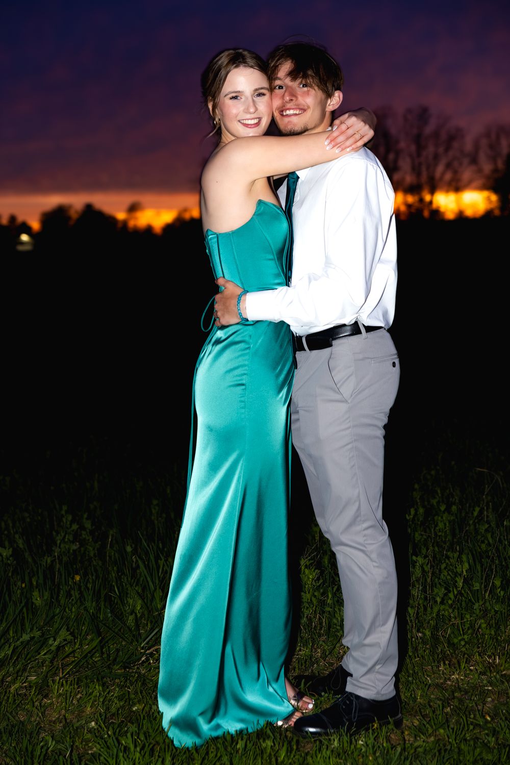 Two figures embrace against a sunset backdrop in a grassy field while wearing turquoise and grey clothing.