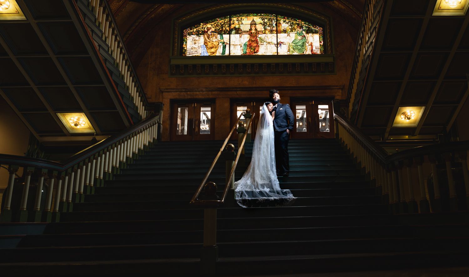 A couple stands on an elegant wooden staircase with stained glass windows in a dimly lit historic building.