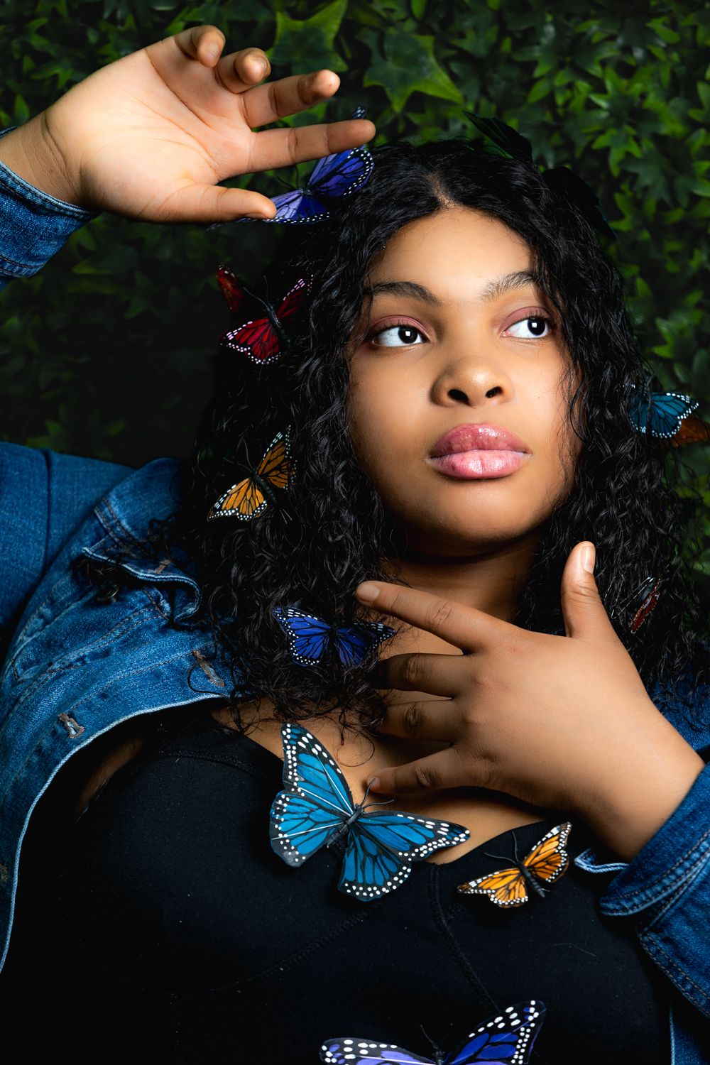 Creative portrait with colorful butterflies adorning black curly hair against green foliage background.