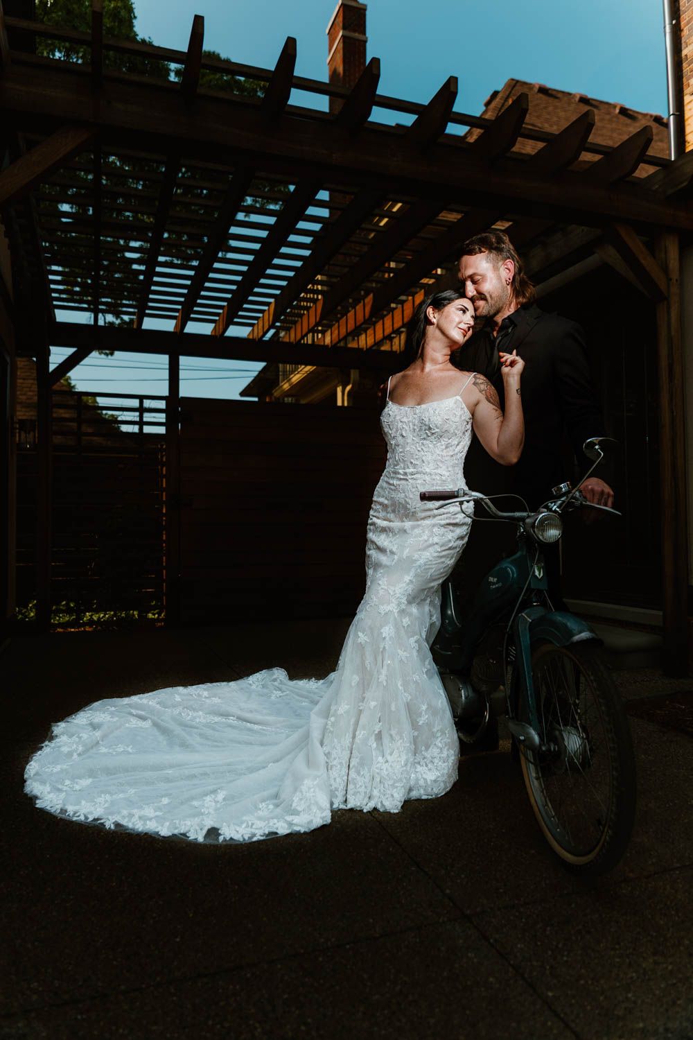 A romantic wedding scene with a stunning lace mermaid gown and vintage bicycle under a wooden pergola at dusk.