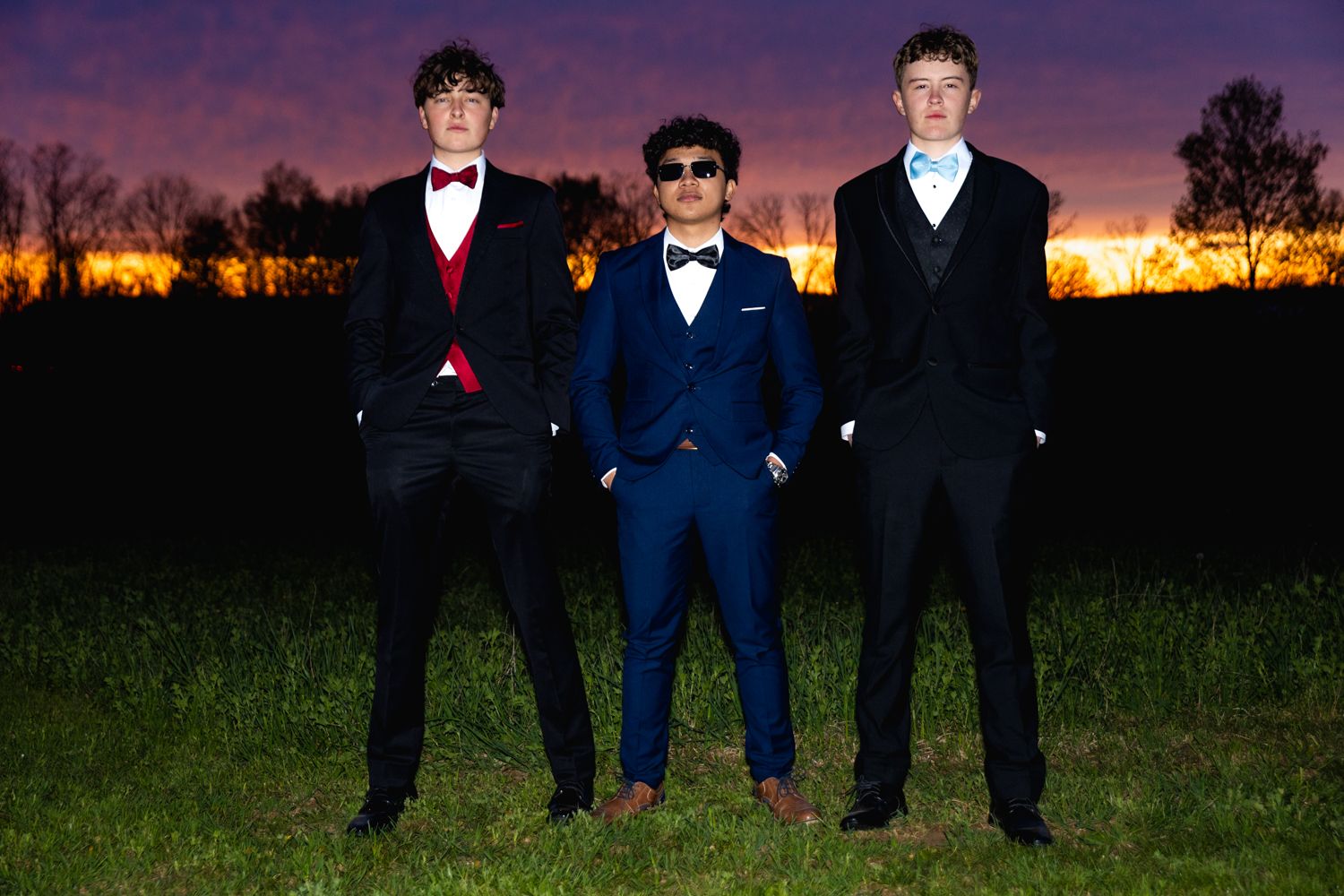 A group in formal attire poses together for a celebratory photo at sunset in a grassy outdoor setting.
