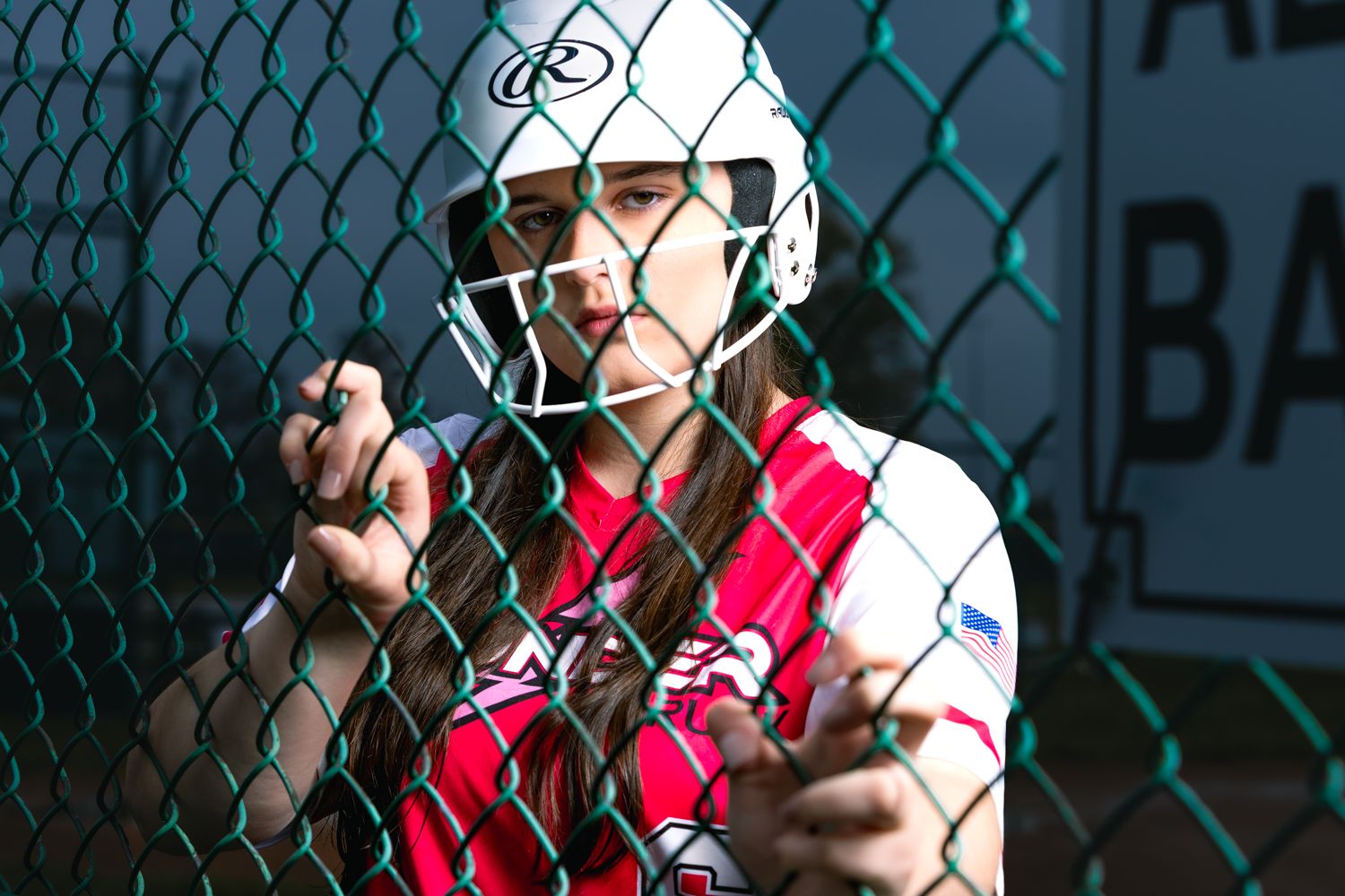 A baseball player in red and white uniform grips a chain link fence while looking through the barrier.