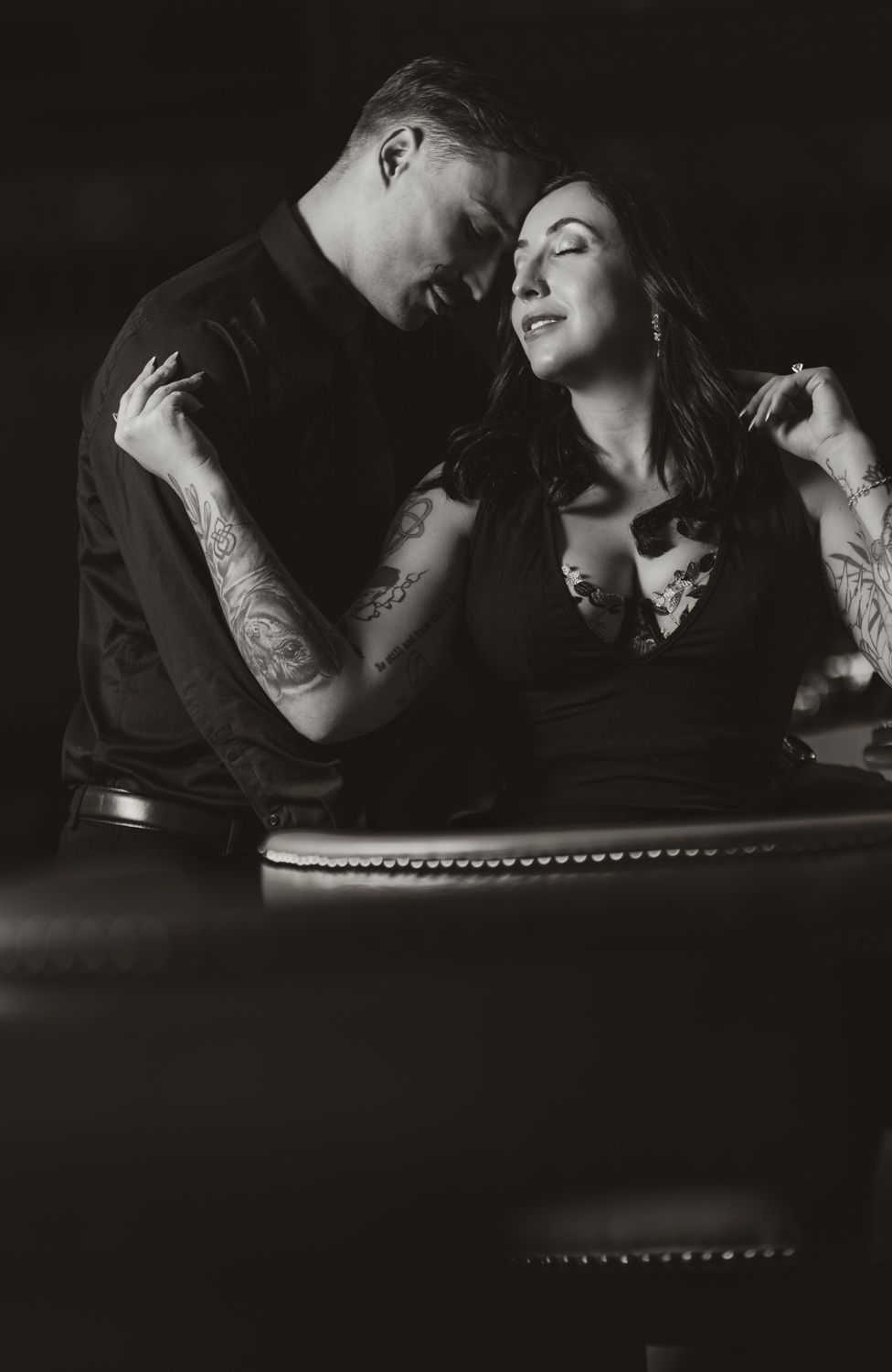 A black and white romantic portrait of a tattooed couple in vintage-style clothing sharing an intimate moment.