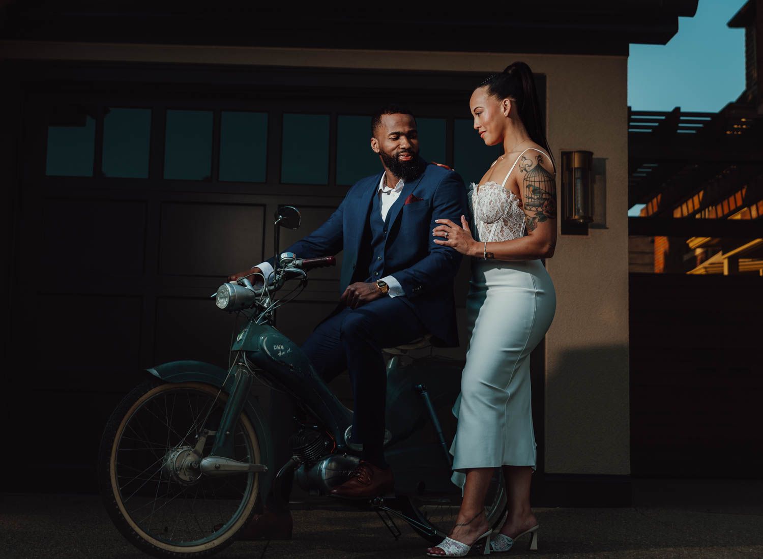 A stylishly dressed couple poses together with a vintage bicycle at dusk against a dark urban backdrop.