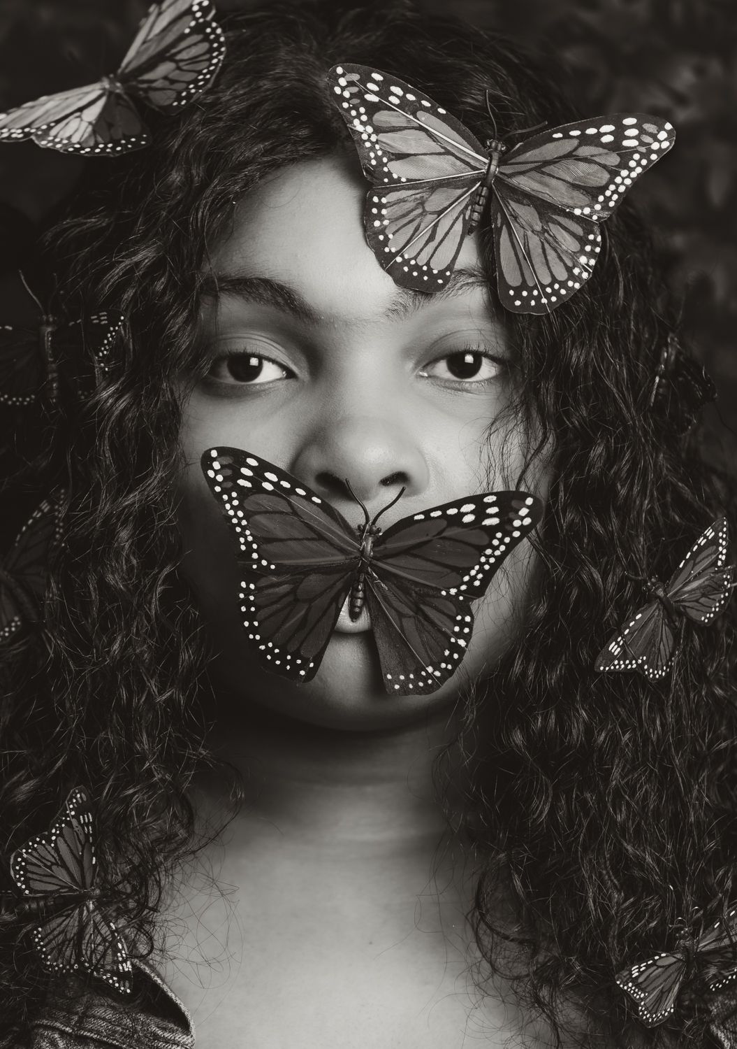 A dreamy black and white portrait with monarch butterflies delicately resting on tousled dark curls in an ethereal composition.
