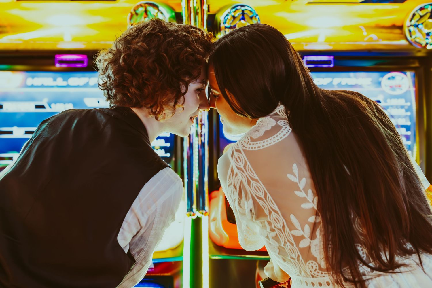 Two people lean in together at an arcade with bright neon lights glowing in the background.
