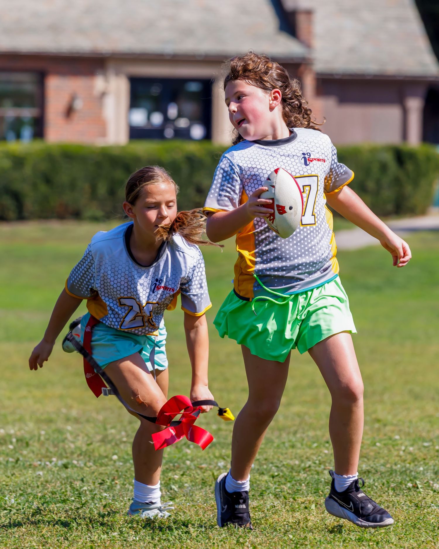 Two girls playing flag football on a grass field, one carrying the ball while the other reaches for her flag.