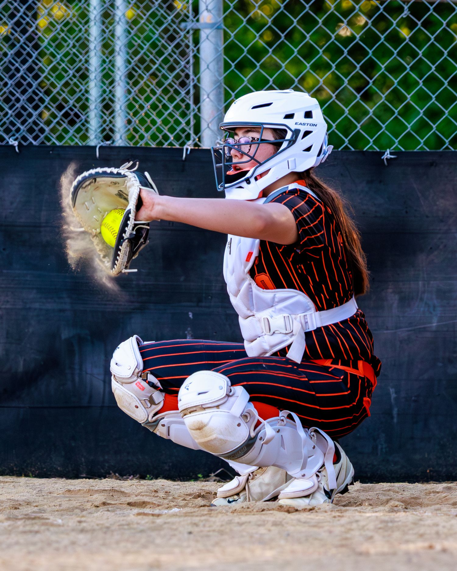 Softball catcher in crouch position catches a yellow ball with glove during a game.