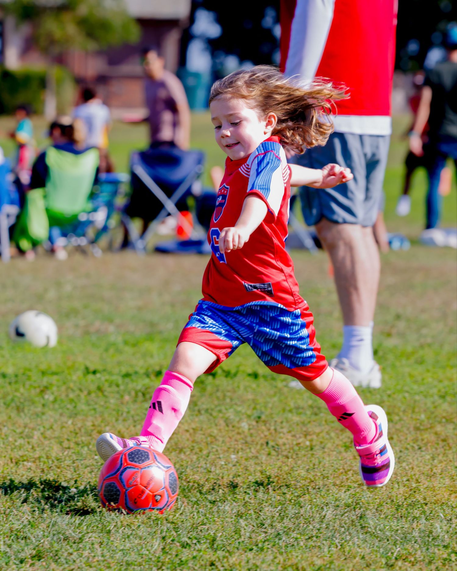 Young soccer player in red jersey and pink socks dribbles ball during outdoor match on grass field.