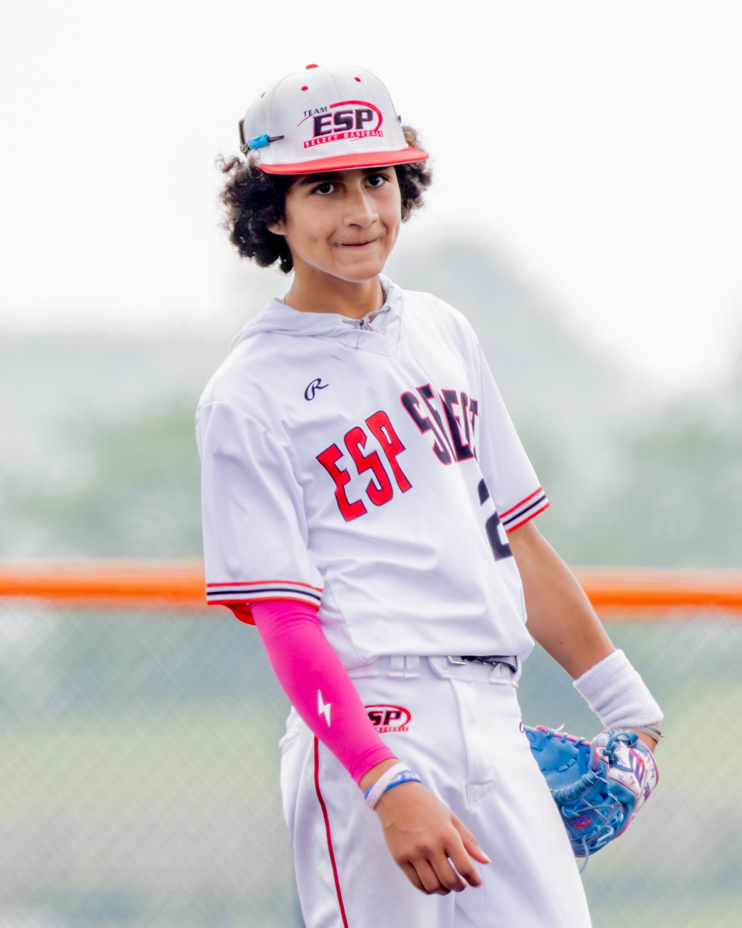 Youth baseball player in white uniform with red accents and pink arm sleeve stands on field during game.