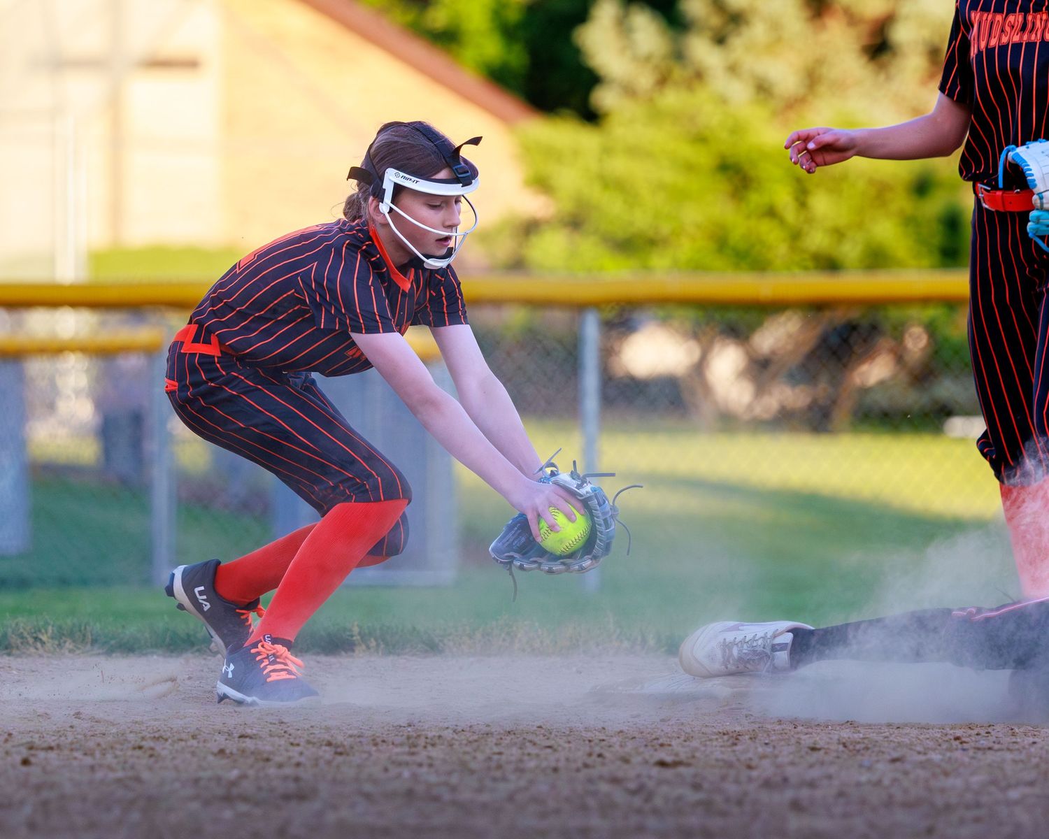 Softball player catches ball at base in cloud of dust during competitive game, wearing black and red uniform with face guard.