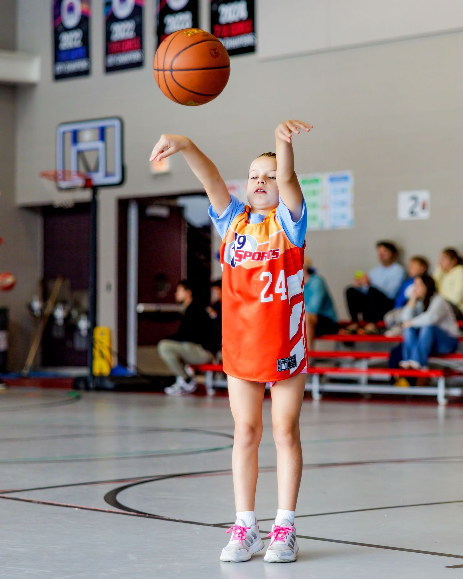 Kids in orange and blue uniforms practicing basketball skills on an indoor court.