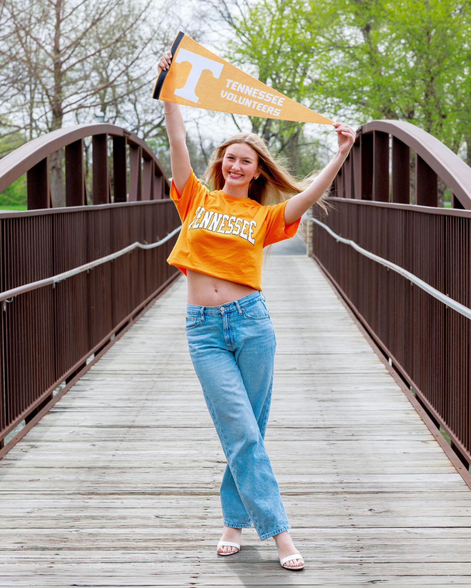 Young woman in orange Tennessee Volunteers crop top holding pennant flag on wooden bridge outdoors.
