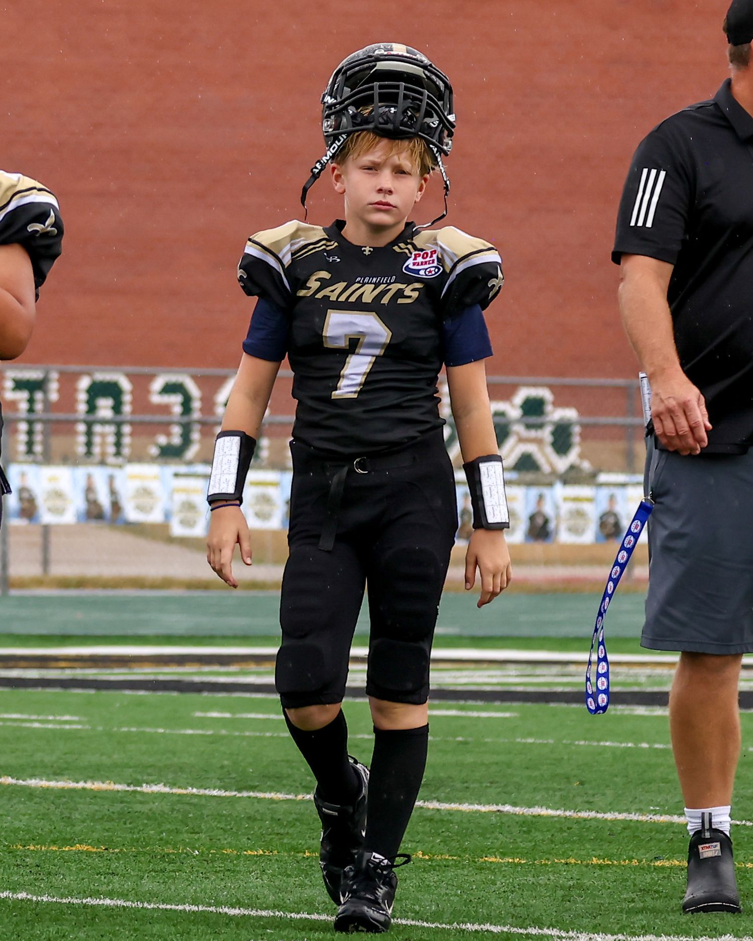 Young football player wearing Saints jersey number 7 stands on field with helmet raised, dressed in black uniform.