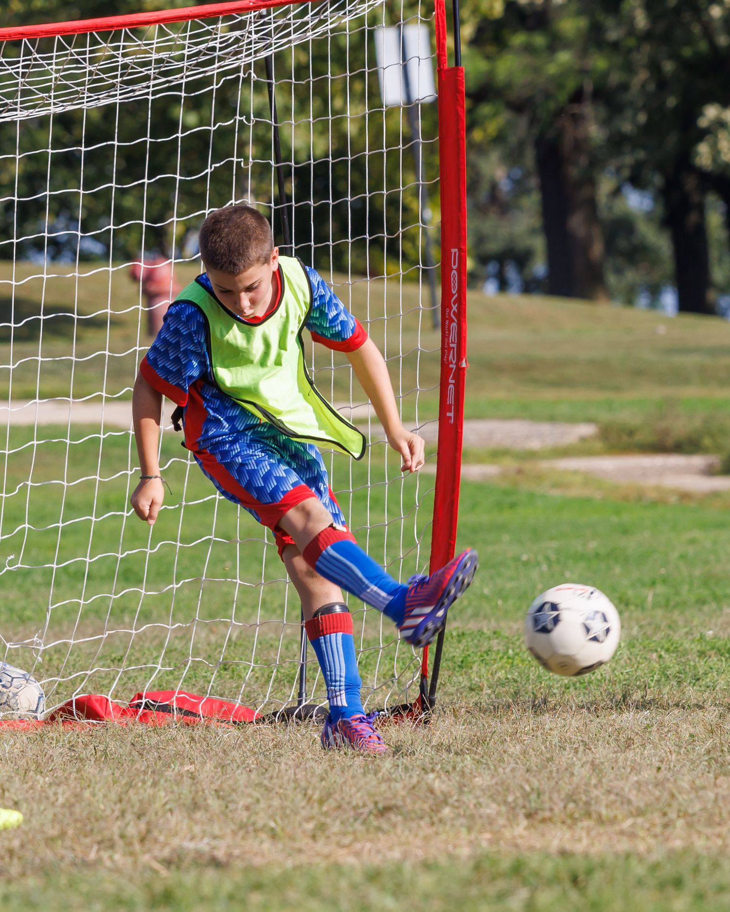 Young boy in yellow vest kicking soccer ball near red goal net on green grass field during youth soccer practice.
