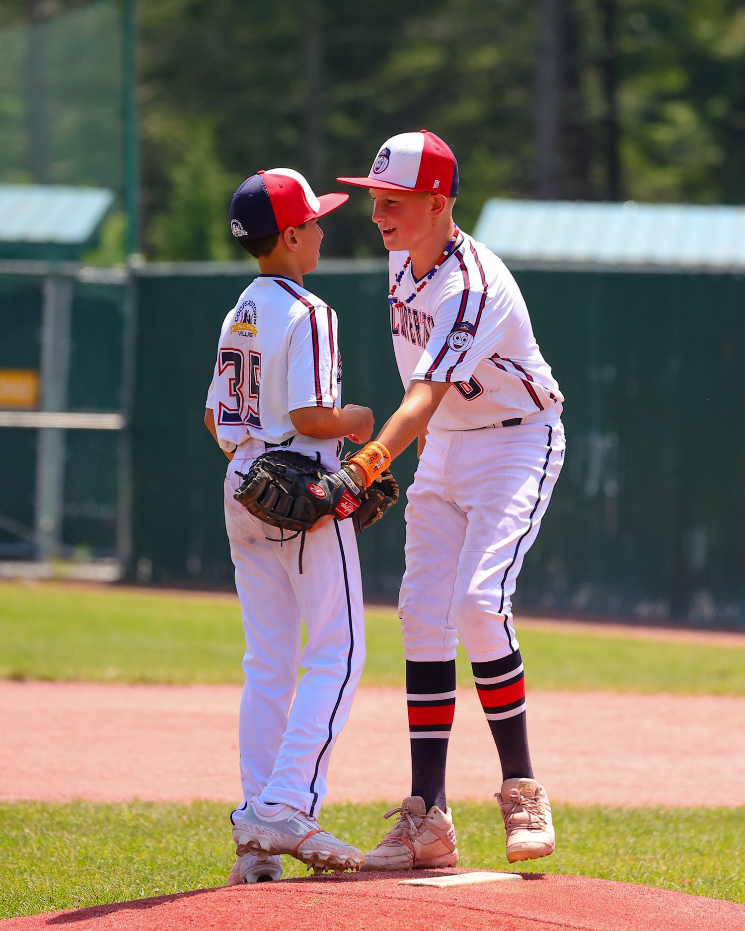 Two youth baseball players in white uniforms kneel down to examine something on the baseball field during a game.