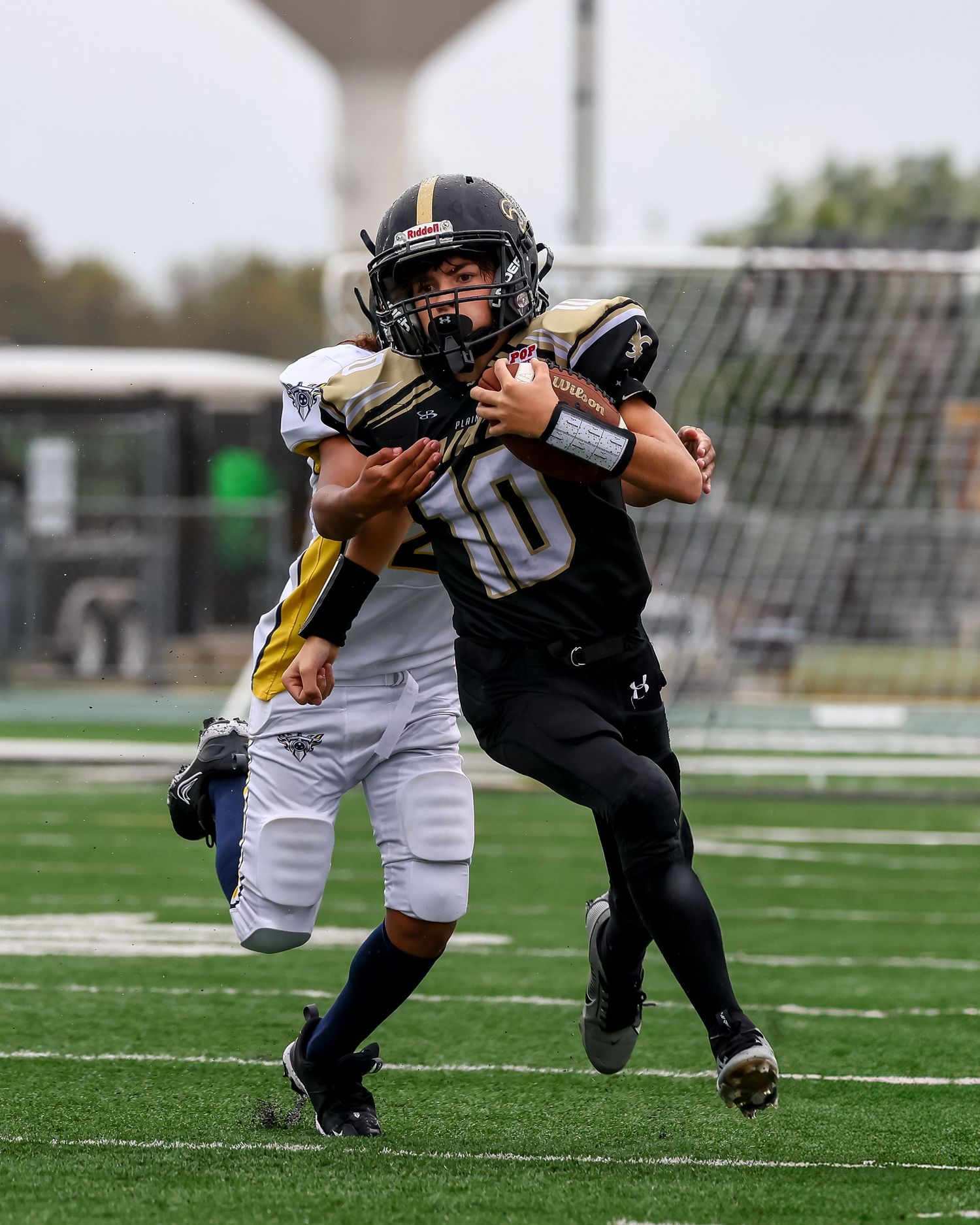 Youth football player in black uniform carrying ball while being tackled by opponent in white during game.