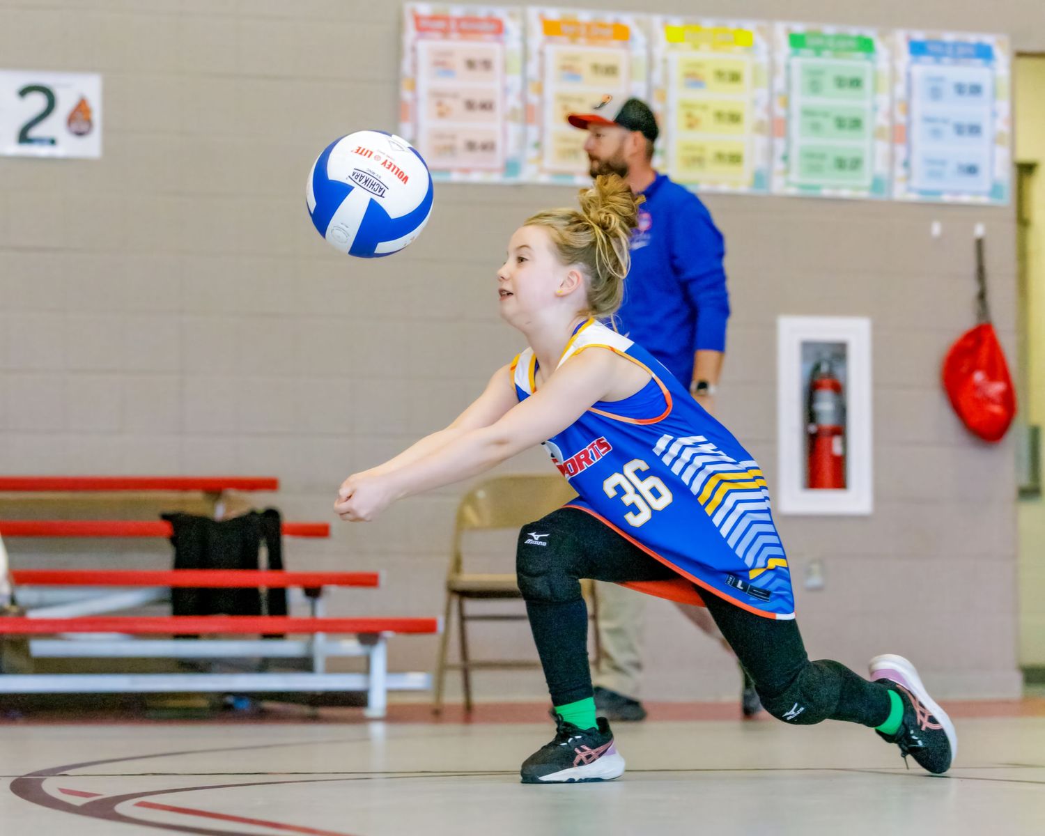 Volleyball player in blue jersey and black pants dives to save ball during indoor match.