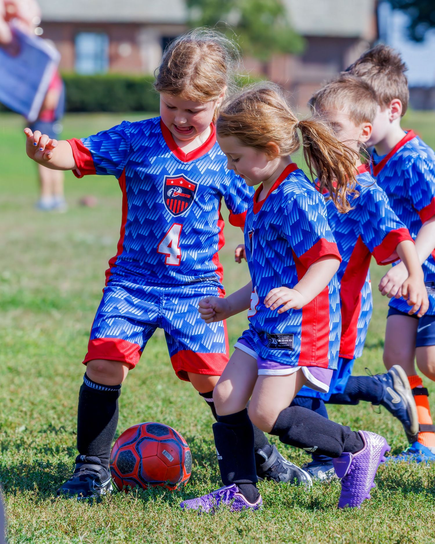 Young children in blue and red soccer uniforms compete for a red ball during a youth soccer game on a green field.