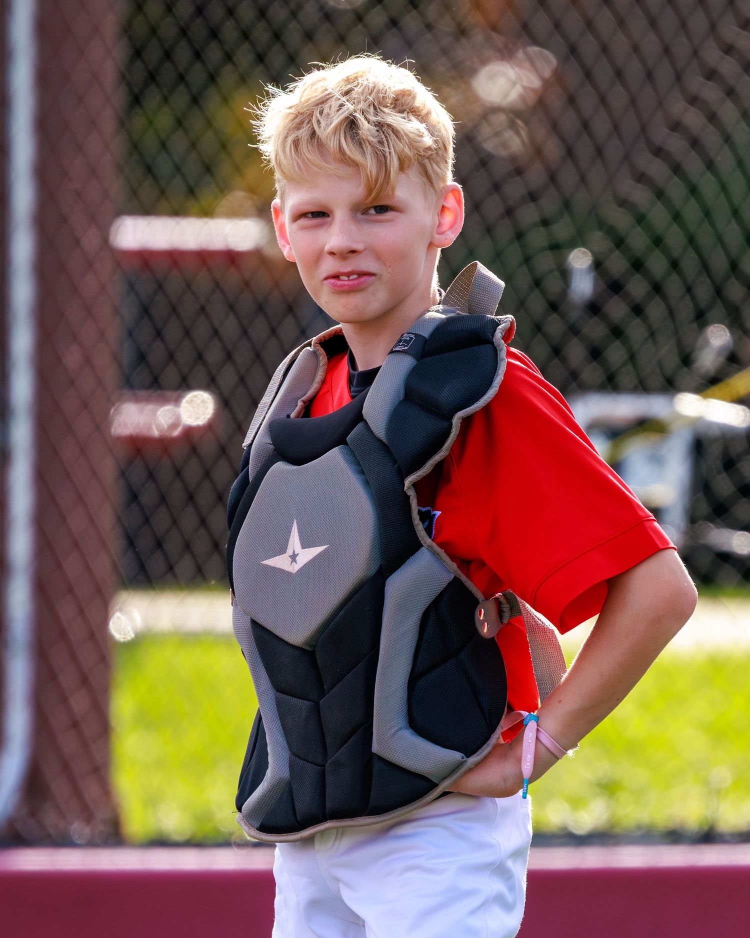 Young baseball catcher wearing chest protector gear in red uniform, posing confidently on baseball field.