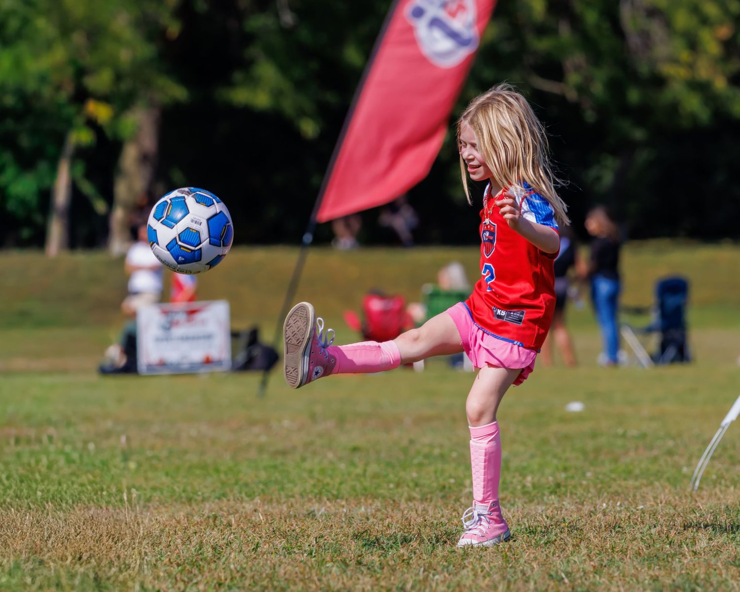 Soccer ball rolling on grass field with red flags and players visible in the background.