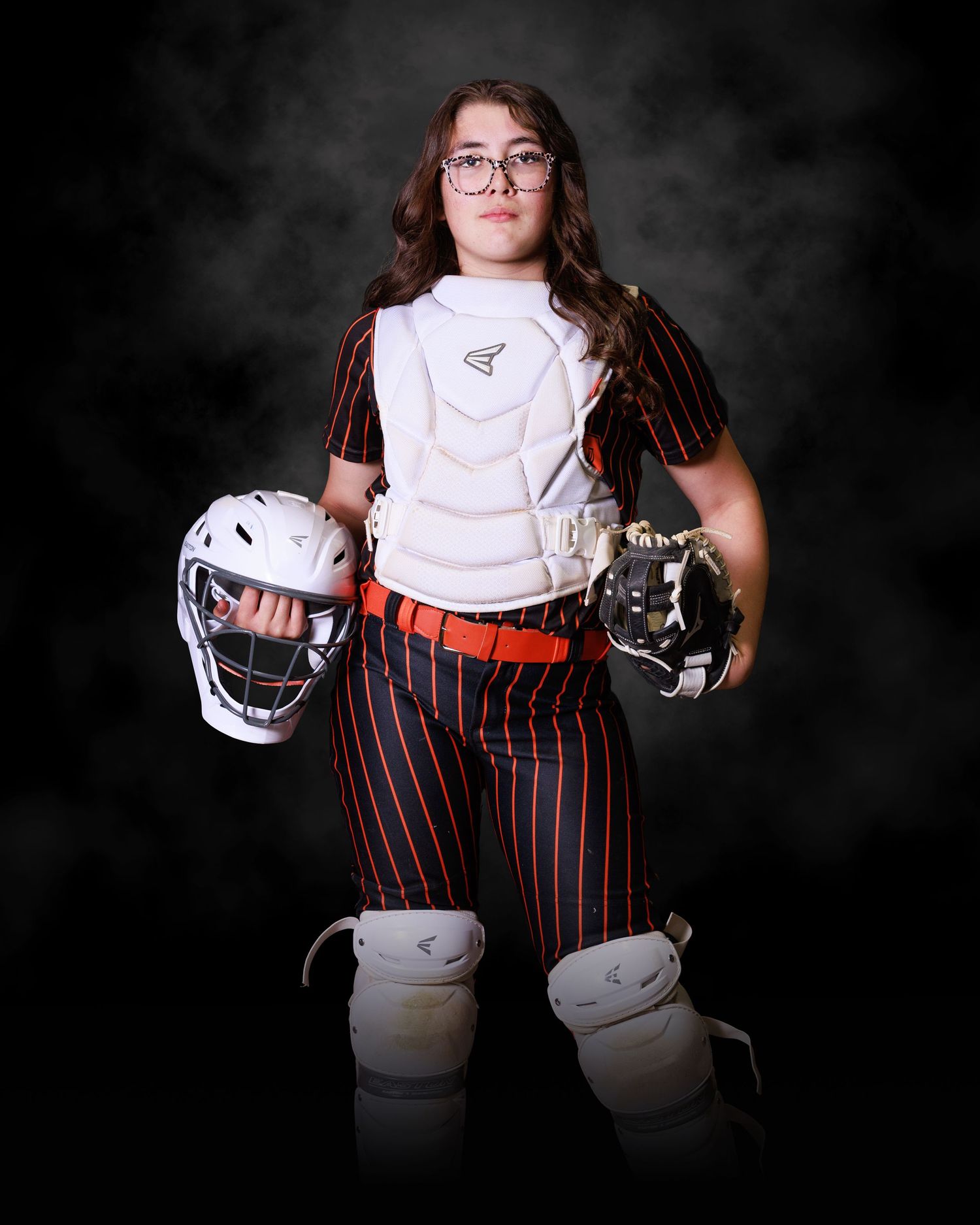 Young softball catcher in pinstripe uniform holding helmet and glove, wearing chest protector and shin guards, posing for team photo.