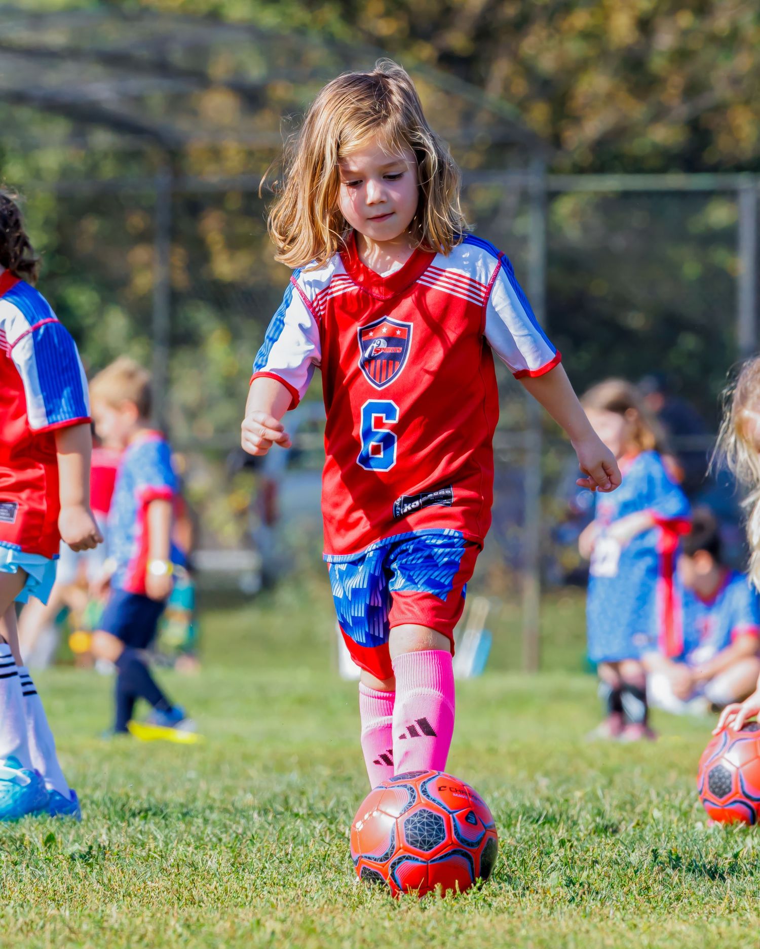Young girl in red number 6 soccer jersey dribbling a ball during a youth soccer game on a sunny day.
