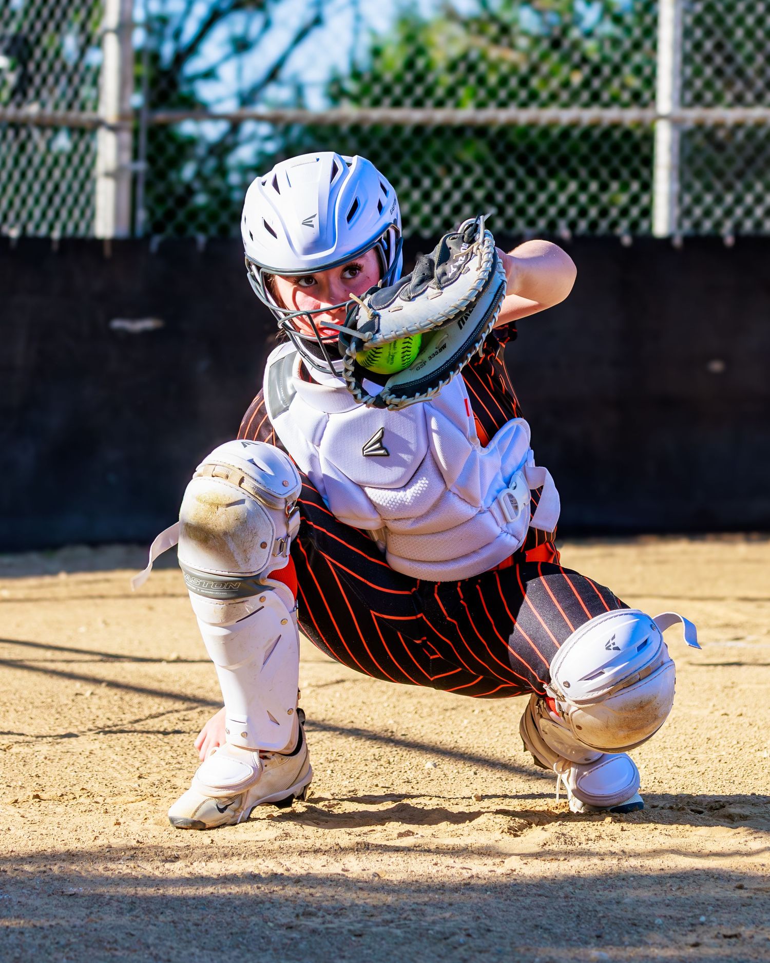 Female softball catcher in white Easton gear crouching in ready position, wearing helmet and chest protector on dirt field.