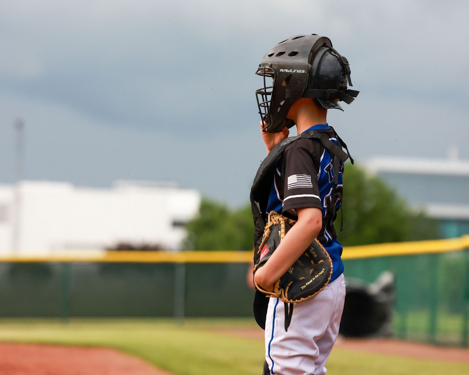 A youth baseball catcher in protective gear stands on the field during a game with a cloudy sky overhead.