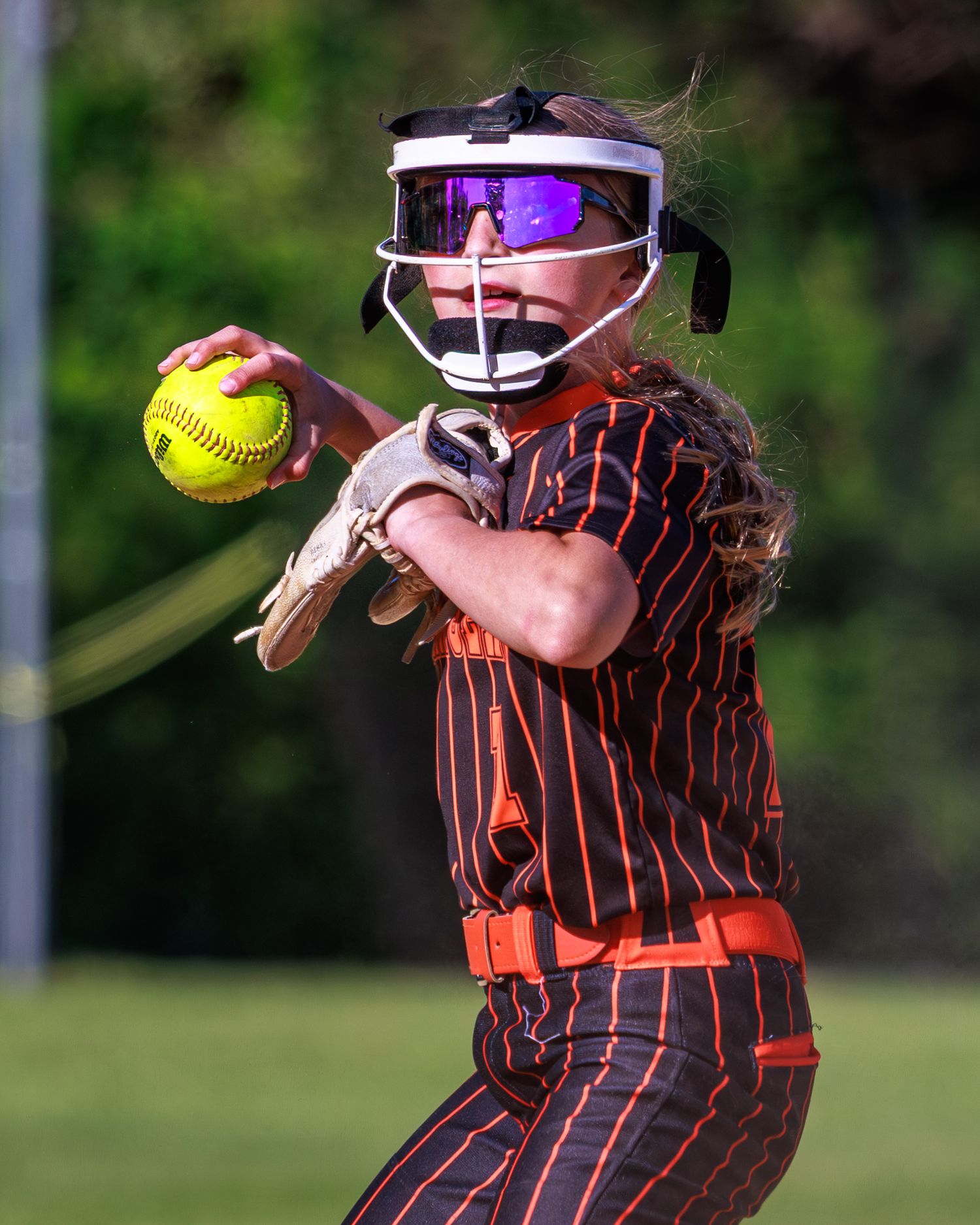 Female softball pitcher in orange and black uniform winding up to throw, wearing protective face mask and holding yellow ball.