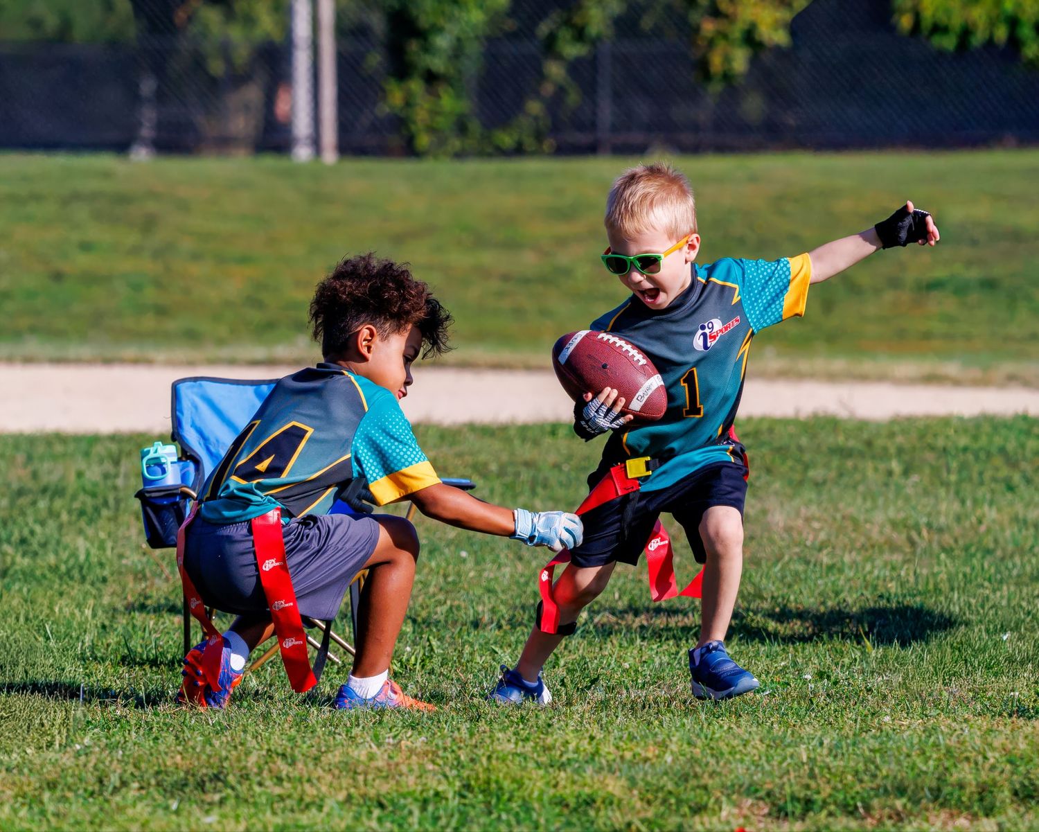 Two young children playing flag football on a green grass field in colorful jerseys during a youth league game.