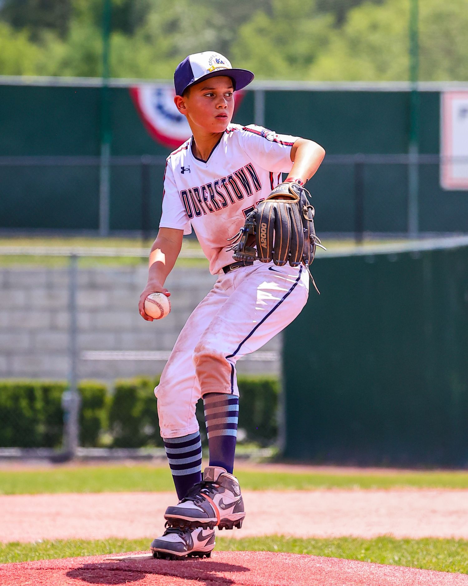 Youth baseball pitcher in white uniform follows through on delivery motion during game on sunny day.