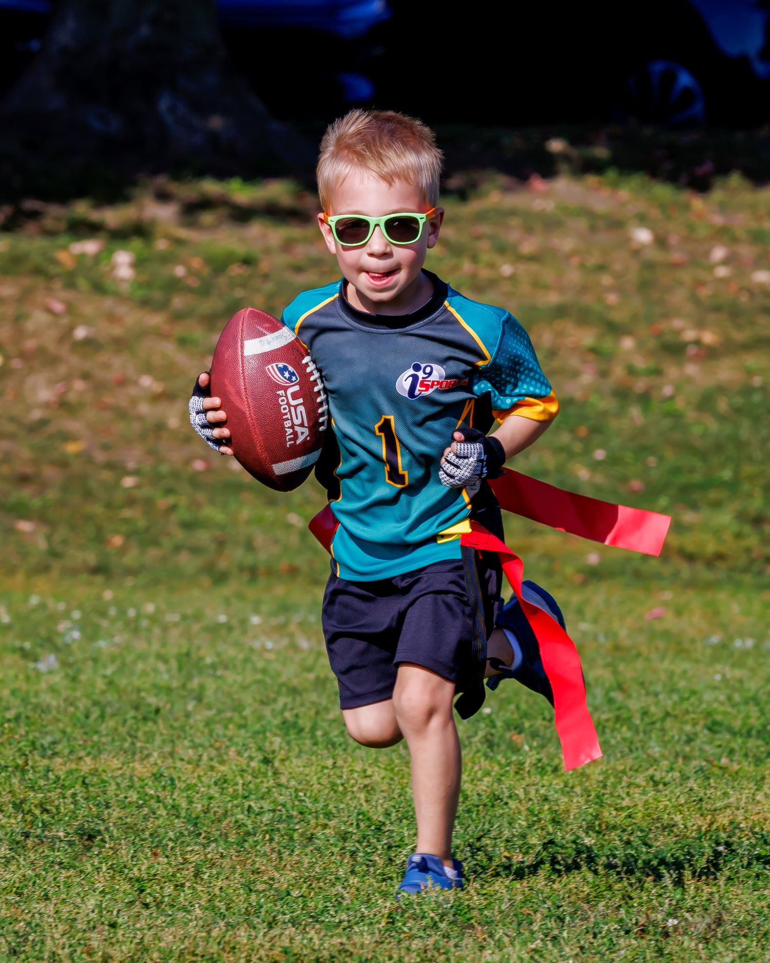 A young player in a teal shirt and navy shorts plays flag football on a grassy field in autumn.