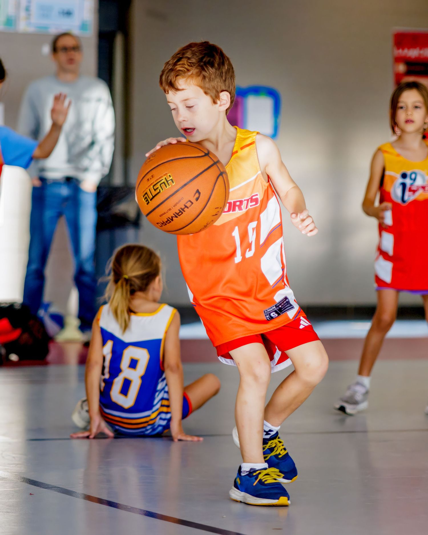 Children practicing basketball drills and dribbling moves in a gym with orange and blue jerseys.