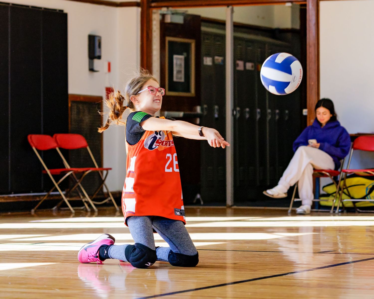 A volleyball player in an orange jersey kneels on the court during a game while a ball floats nearby.