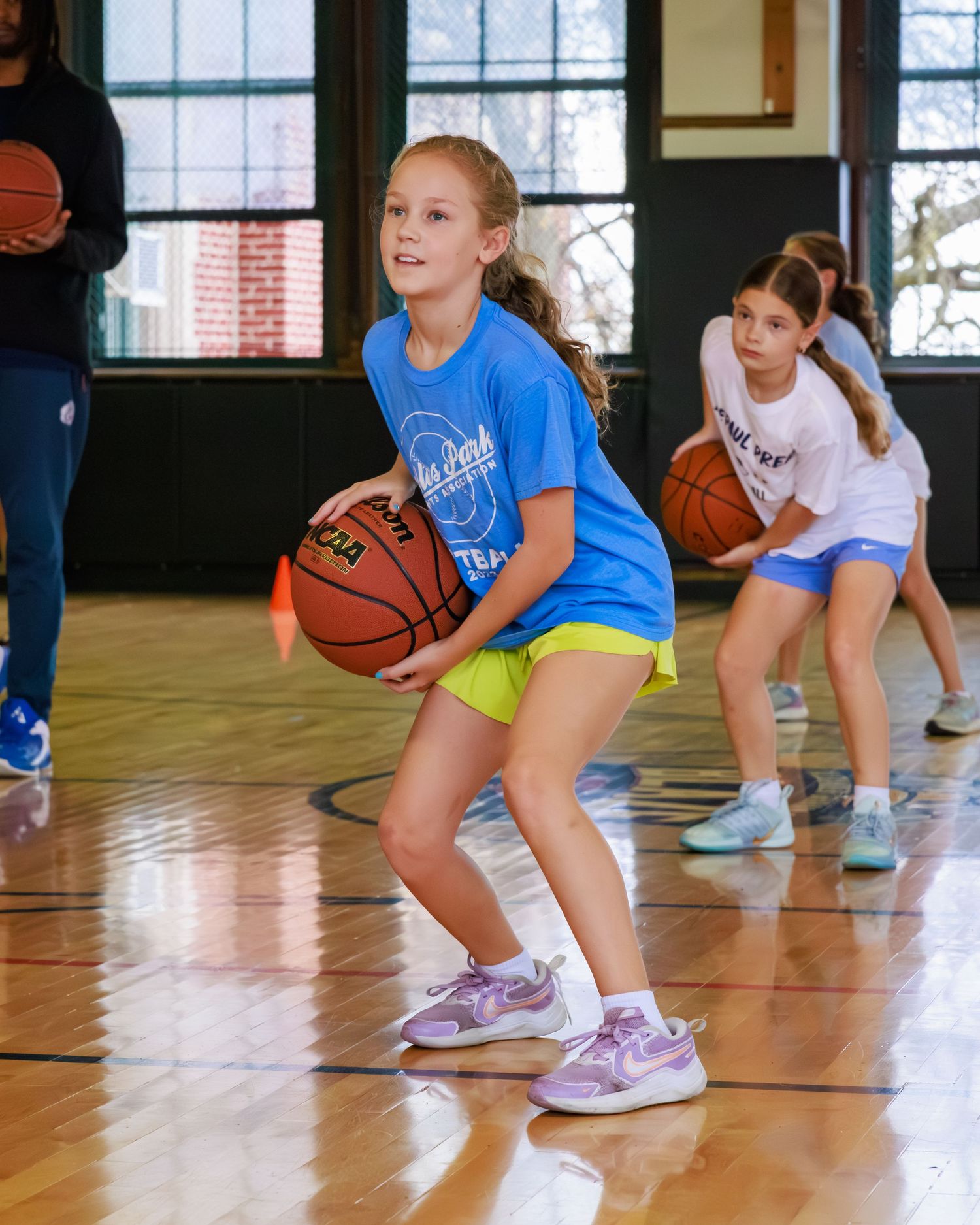 Young athletes practice basketball drills in a row on a gymnasium floor while wearing blue and white uniforms.