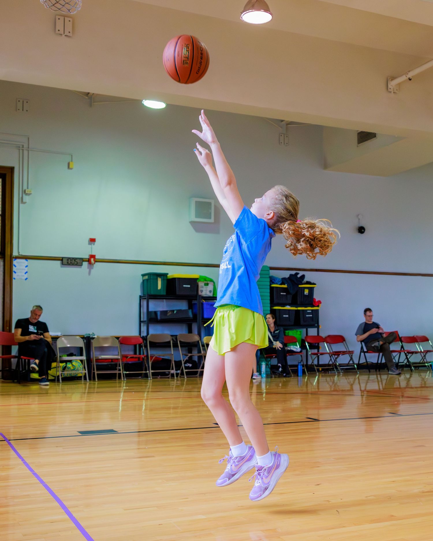 A person in blue and yellow athletic wear jumps while playing basketball in a gymnasium with chairs along the wall.