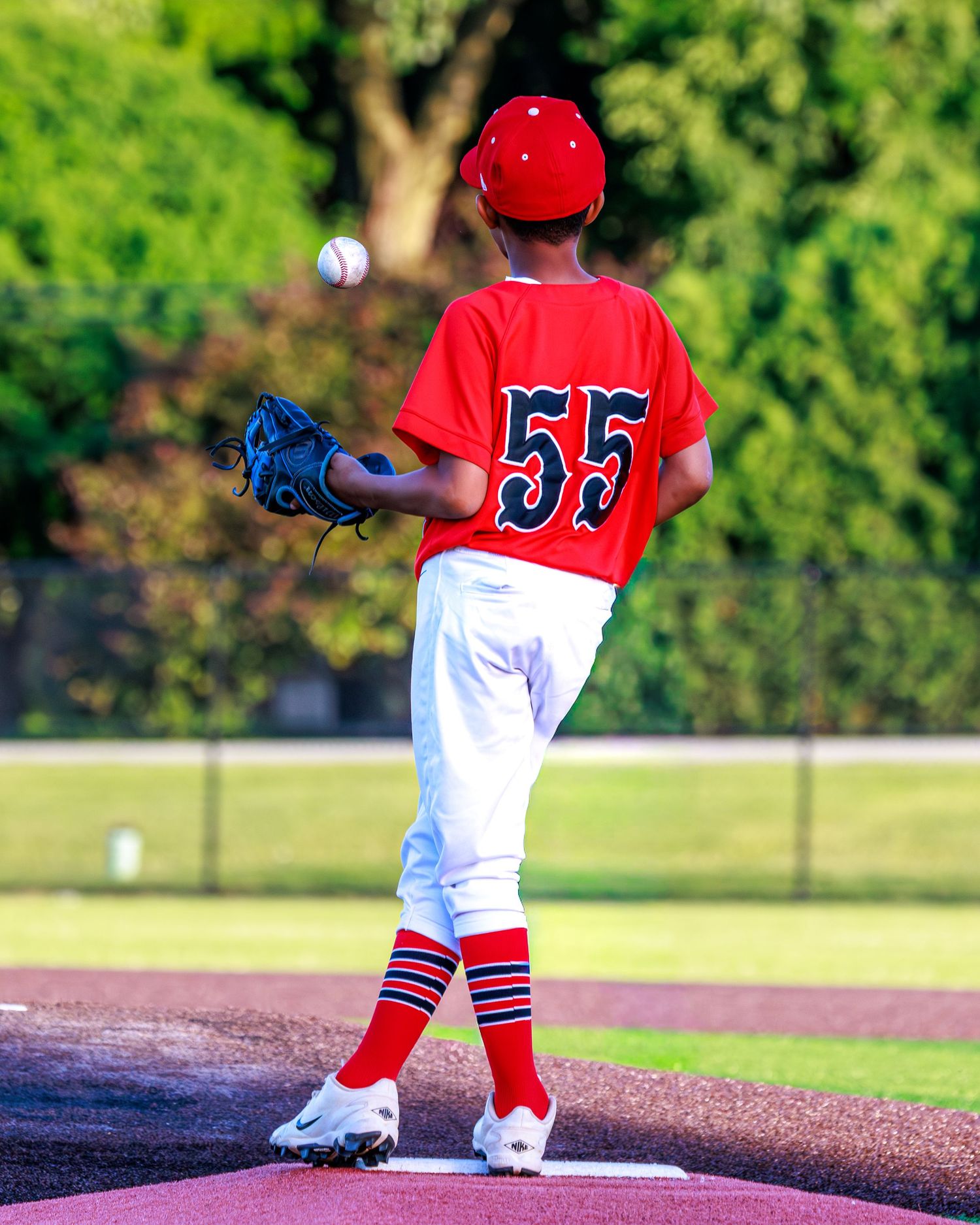 Young baseball pitcher #55 in red uniform winds up on mound, glove raised, ready to throw.