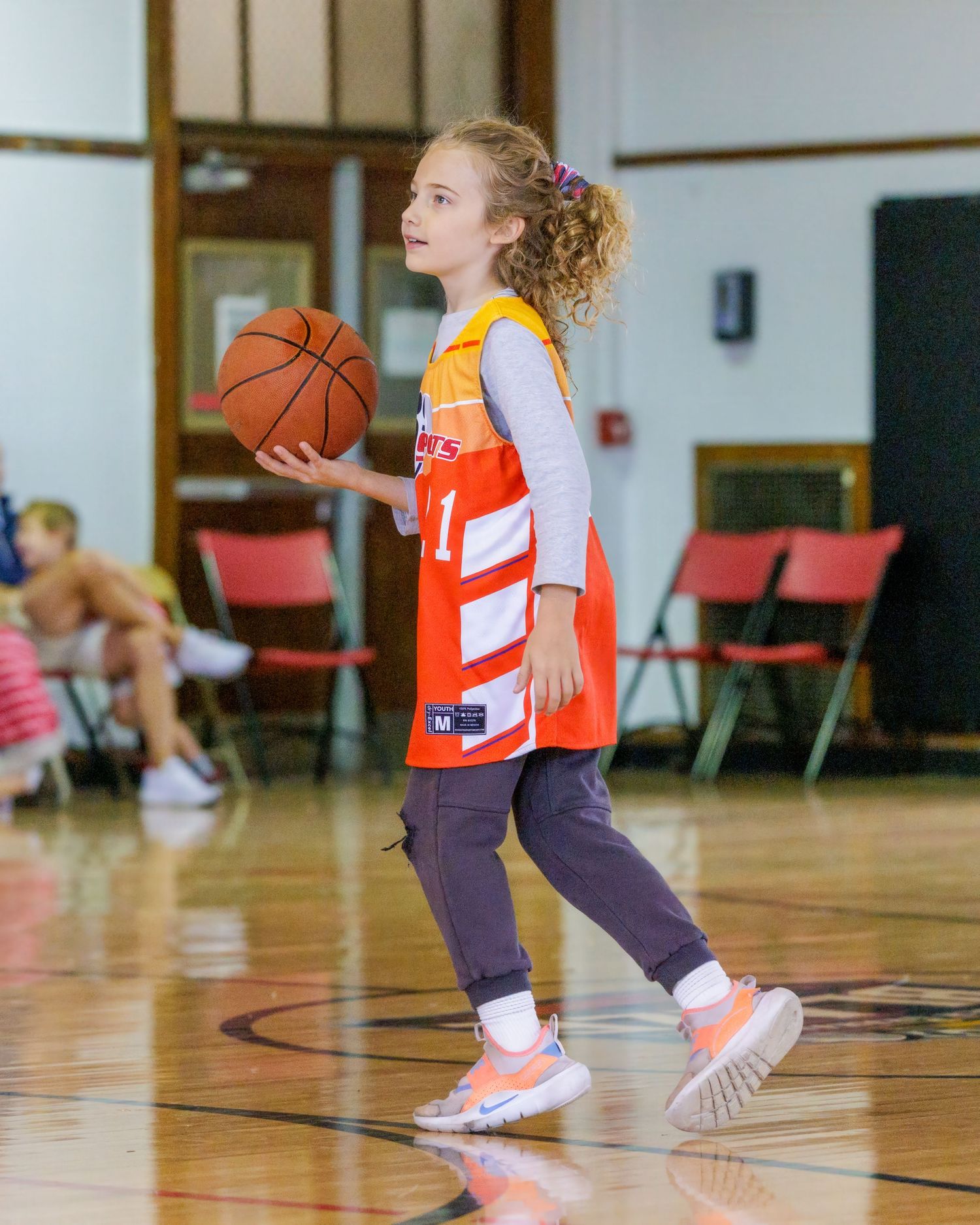 Young girl in orange basketball jersey dribbling ball on indoor court during youth league game.