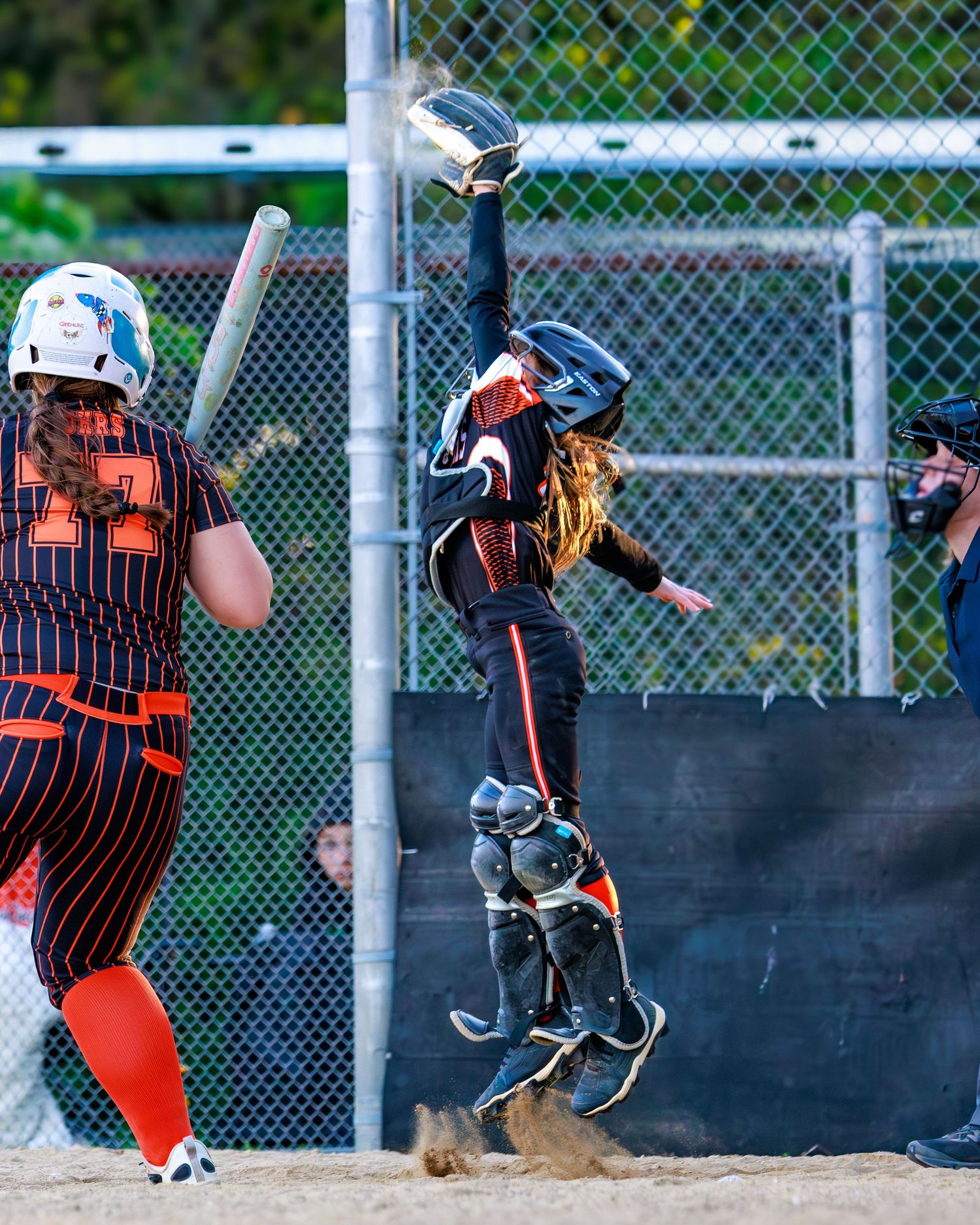 Softball catcher leaps high to catch a ball during a game while batter stands ready at home plate.