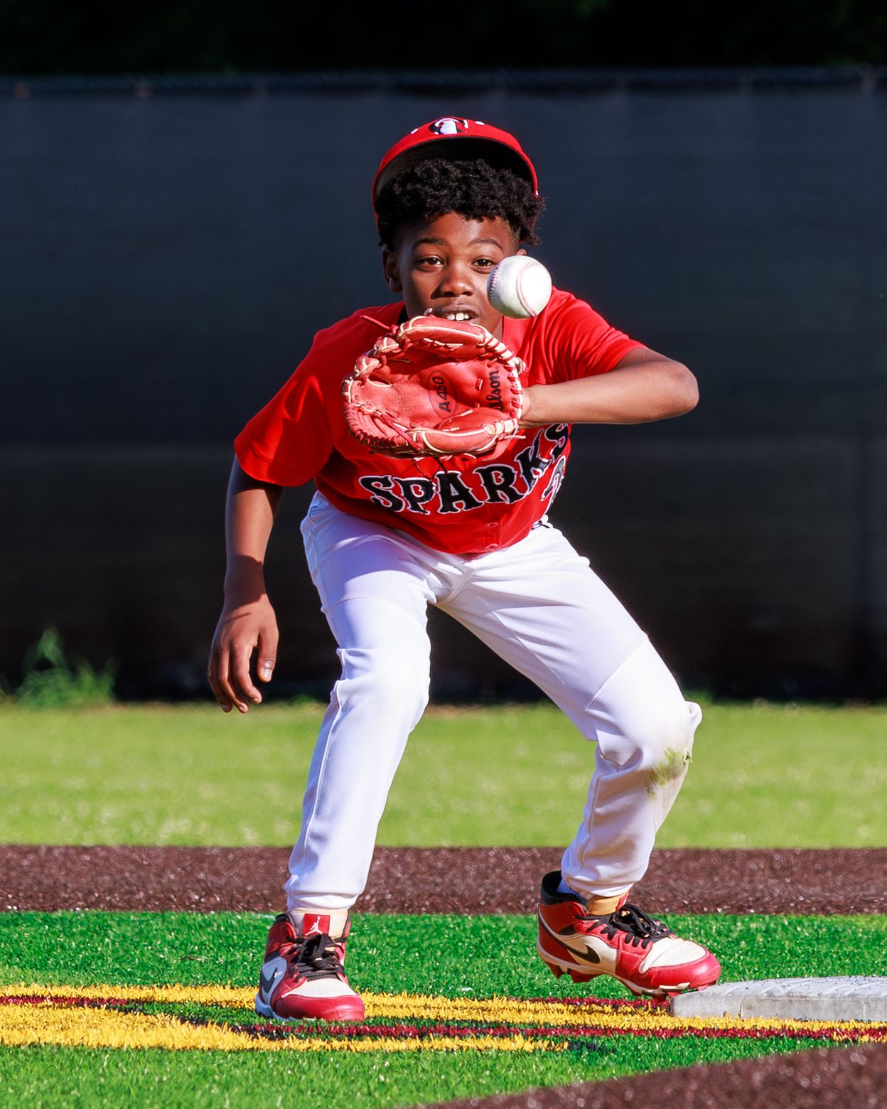 Young baseball player in red Sparks uniform catches a ball with mitt while fielding at base on green turf.