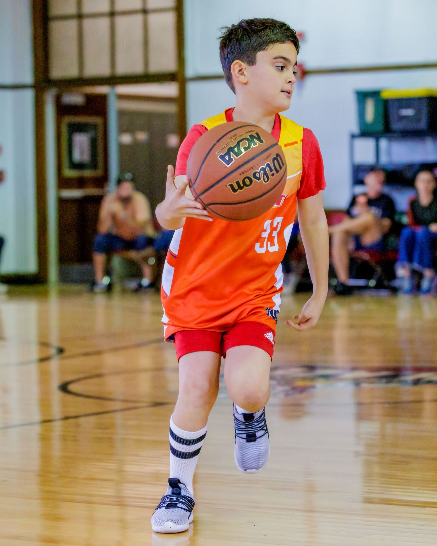 Young boy in orange jersey #33 dribbling a Wilson NCAA basketball on an indoor court during a youth game.