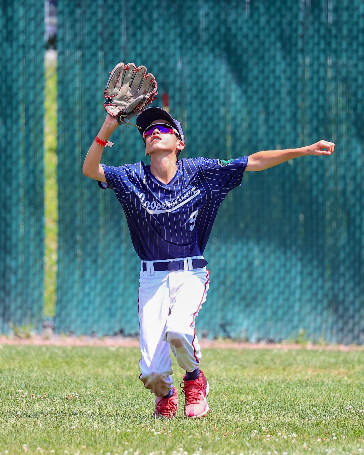 Young baseball player in navy uniform falling backwards while attempting to catch a ball in the outfield.
