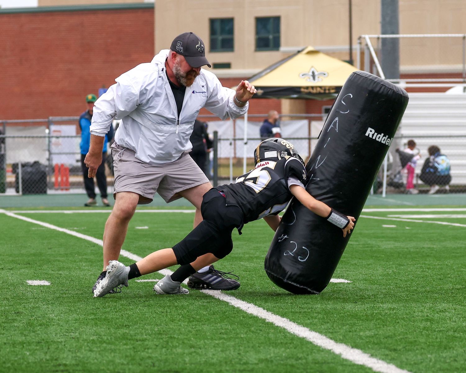 Football coach instructs young player hitting a Riddell blocking dummy during practice drill on turf field.