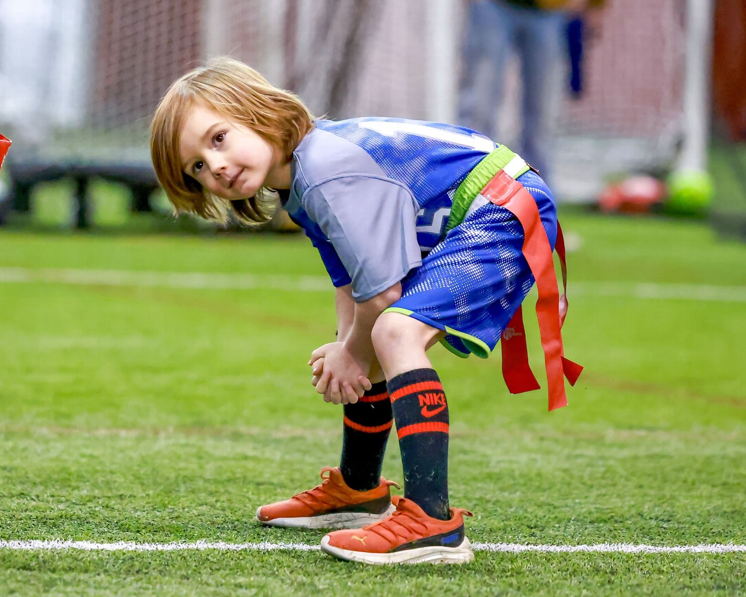 A young figure in blue clothing and red striped socks bends over during an indoor game on green turf.