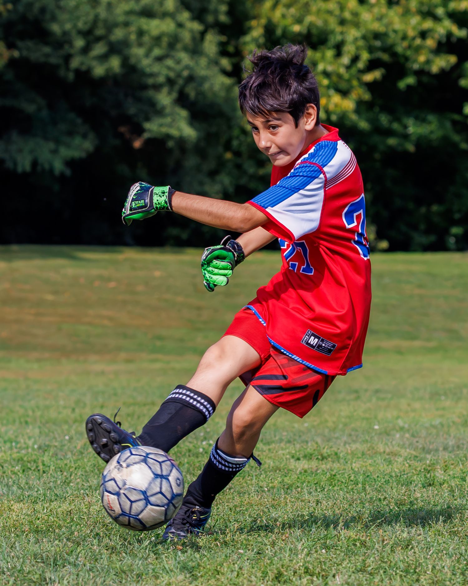 Young boy in red soccer jersey and green goalkeeper gloves kicking a soccer ball on a green grass field.