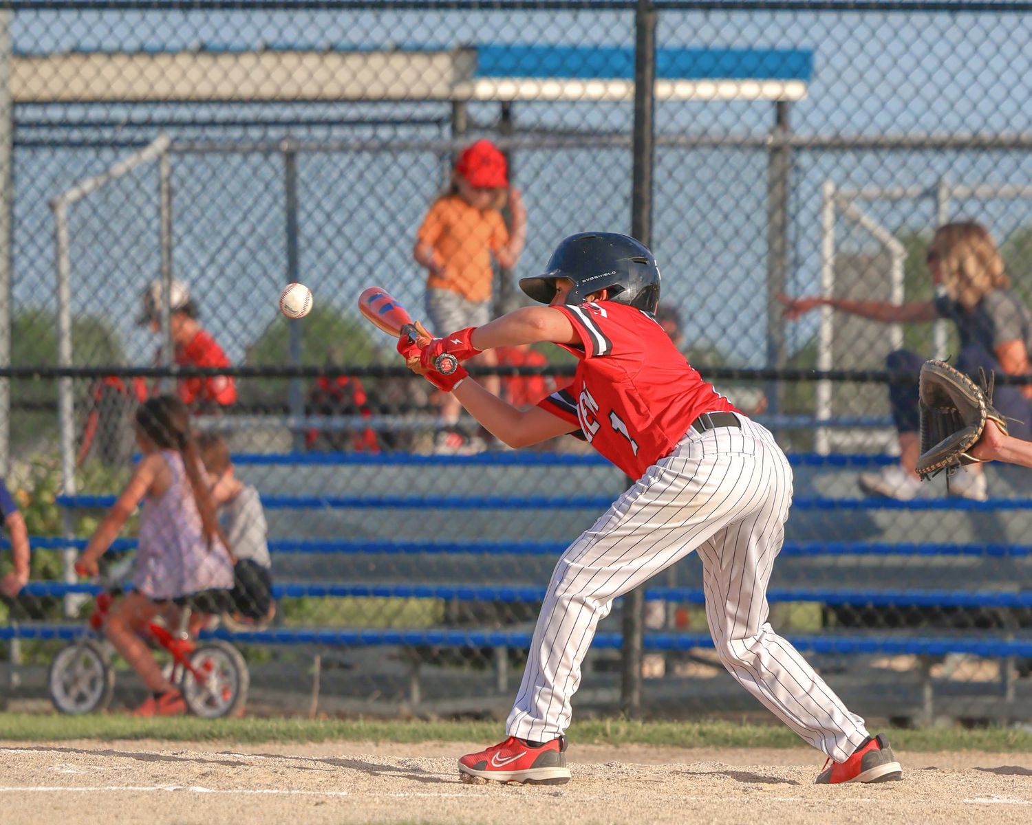 Baseball batter watches a pitch approach while positioned in a ready stance near home plate during a sunny afternoon game.