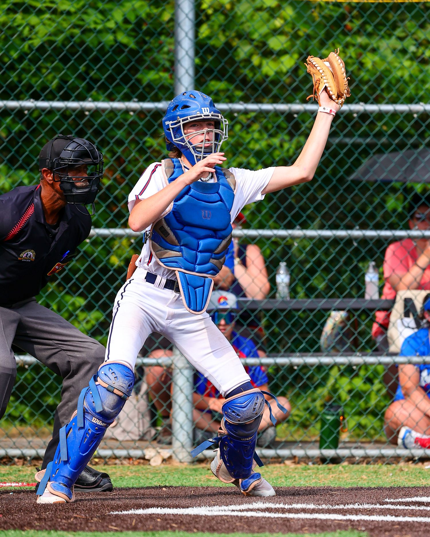 Baseball catcher in blue uniform reaches up to catch a ball during game with chain link fence and trees in background.