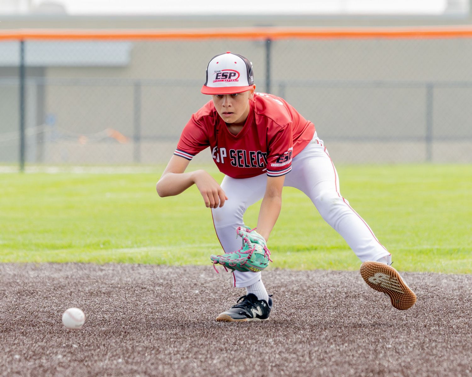 Baseball player in red uniform crouches to field a ground ball during infield practice on dirt field.