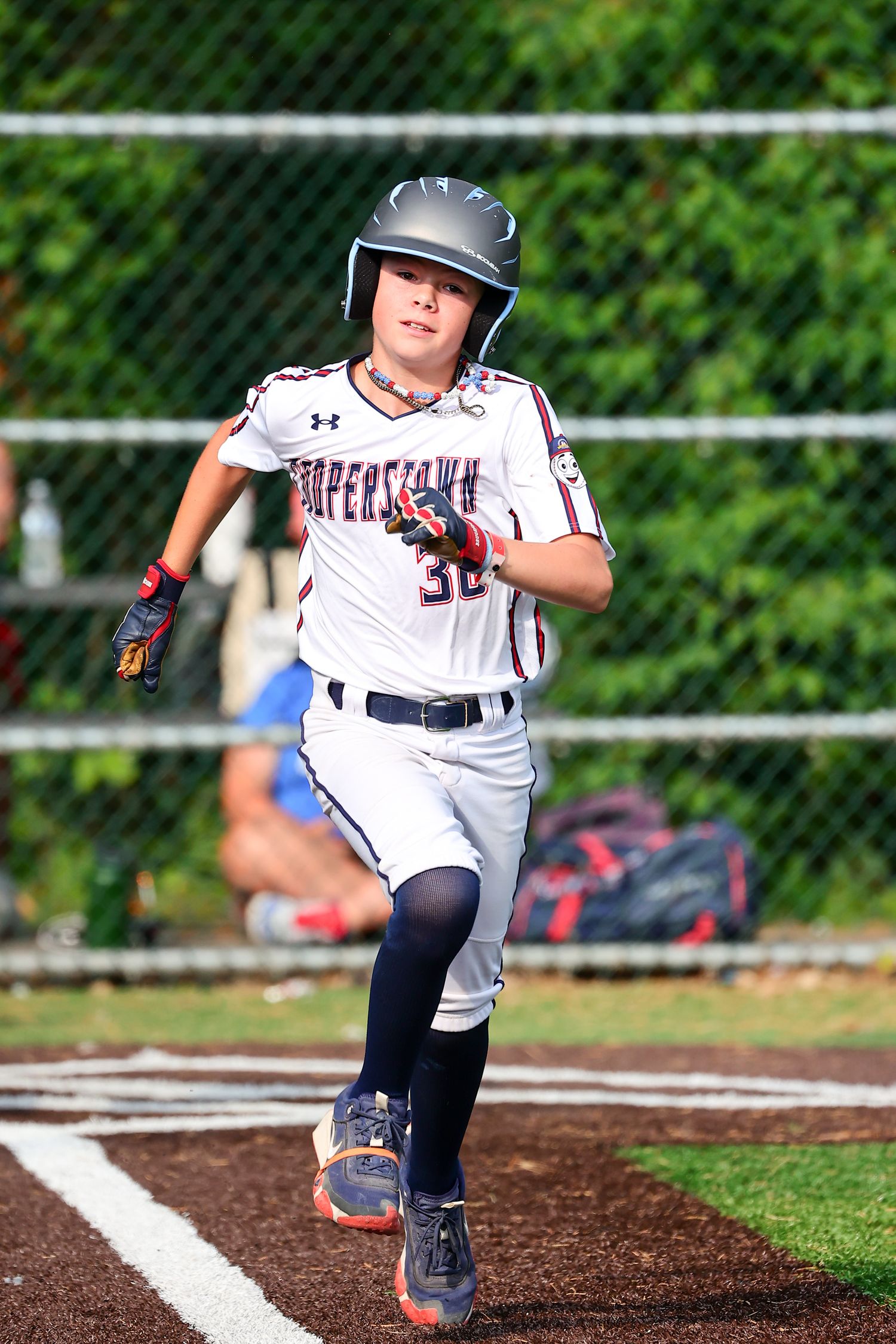 Baseball player in white uniform and helmet runs on dirt path during game with green fence in background.